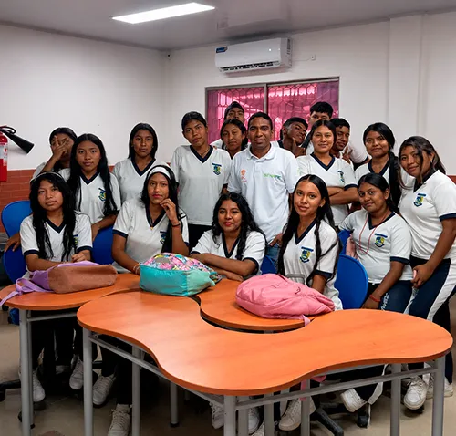 Grupo de estudiantes posa junto a su instructor de SENATIC en un salón de clases. Los jóvenes, vestidos con uniforme blanco y azul, rodean una mesa mientras sonríen a la cámara. El instructor, ubicado en el centro del grupo, lleva una camiseta blanca con los logotipos institucionales de SENATIC. El aula es amplia, iluminada y cuenta con aire acondicionado, ventanas laterales, sillas azules y mesas de trabajo. La imagen transmite un ambiente de aprendizaje, compañerismo y formación en competencias tecnológicas.