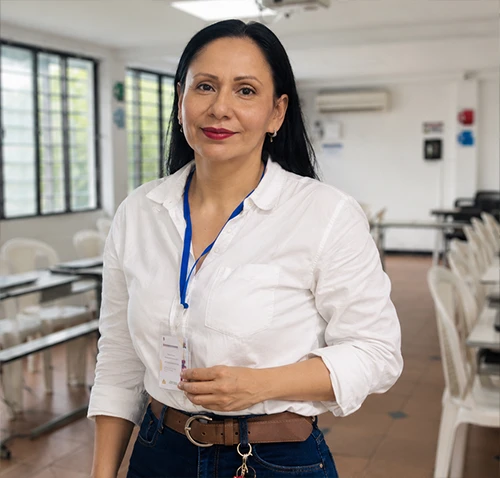 Mujer de cabello oscuro y camisa blanca de manga larga, de pie frente a la cámara en un aula de informática