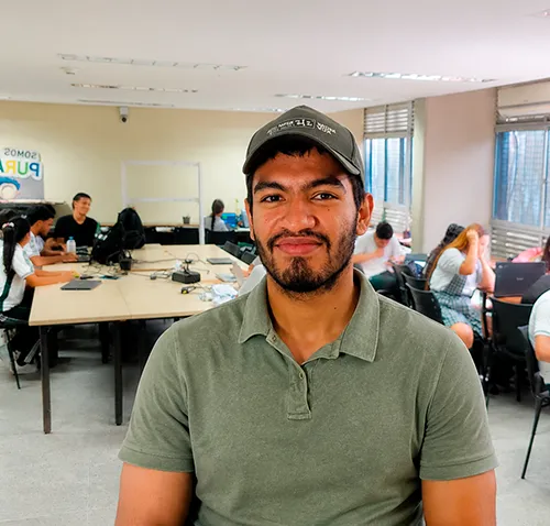 Un instructor de SENATIC posa frente a la cámara en un aula de informática mientras, al fondo, varios estudiantes trabajan en equipos con computadores portátiles