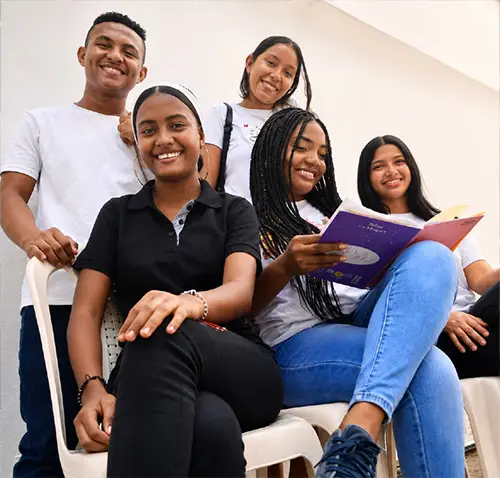Cinco jóvenes sonríen a la cámara mientras comparten un momento de lectura y conversación. Tres mujeres están sentadas en sillas plásticas; una de ellas sostiene un cuaderno abierto, mientras otra mira directamente a la cámara. Detrás de ellas, un joven y una mujer joven permanecen de pie sonriendo. El grupo está en un ambiente tranquilo y luminoso que transmite cercanía, participación y trabajo en equipo.