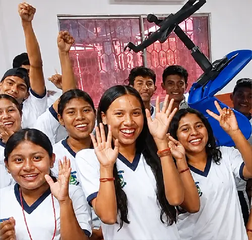 Un grupo de estudiantes de secundaria desde el departamento de La Guajira, con uniforme blanco y azul, sonríe y saluda con entusiasmo dentro de un salón de clases. Varias jóvenes levantan las manos y hacen el gesto de la paz, mientras otros estudiantes aparecen al fondo con expresiones alegres. El aula cuenta con mesas, sillas azules y maletas sobre los pupitres. La imagen transmite un ambiente de compañerismo, participación y entusiasmo durante una actividad escolar.