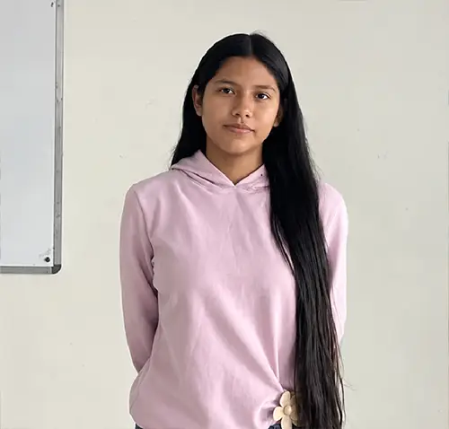 Una estudiante posa de pie frente a la cámara en un salón de clases. Viste una sudadera con capucha de color rosa claro y pantalón de mezclilla azul