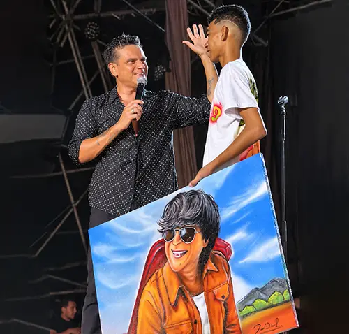 Un artista (Silvestre Dangond), vestido con una camisa negra, sostiene un micrófono mientras sonríe y conversa con un joven que viste una camiseta blanca